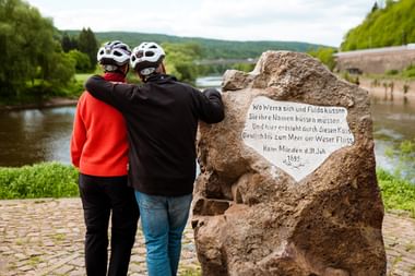 Zwei Radfahrer mit Helmen lesen ein Steinmonument mit deutscher Inschrift neben der Weser in Hann. Münden, mit grünen Hügeln im Hintergrund.