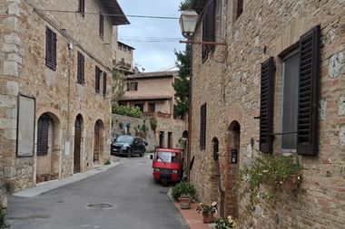 Schmale Steinstraße in San Gimignano mit mittelalterlichen Gebäuden, Holzläden, einem roten Dreirad und Topfpflanzen am Gehweg.