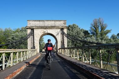 Radfahrerin fährt über eine historische steinerne Hängebrücke am Rhône-Radweg unter blauem Himmel mit grünen Bäumen im Hintergrund.