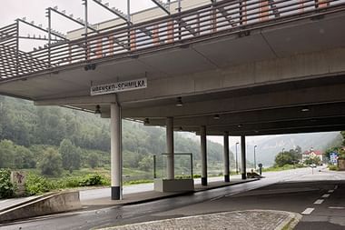Moderne Betonbrücke mit Schild 'Hřensko-Schmilka' über einer Straße. Grüne Hügel und Fluss im Hintergrund unter bewölktem Himmel.