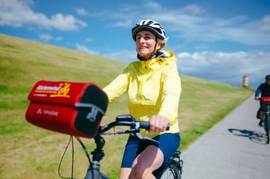 Radfahrerin in gelber Jacke und Helm auf Deichweg mit roten Packtaschen, grüne Grashänge und Leuchtturm im Hintergrund sichtbar.