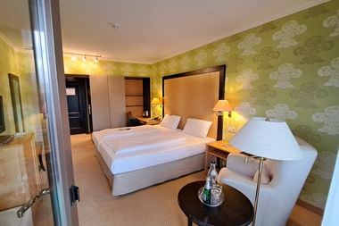 Modernes Hotelzimmer mit Doppelbett, grüner Blumentapete, weißem Sessel, TV und Holzschrank. Warme Beleuchtung schafft gemütliche Atmosphäre.