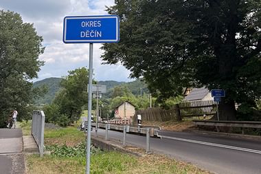 Blaues Ortsschild mit der Aufschrift 'OKRES DĚČÍN' an einer Straße mit Bäumen, Hügeln und einem Fußweg im Hintergrund.
