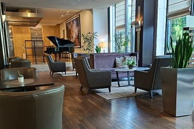 Moderne Hotellobby im Sheraton Essen mit dunklen Sesseln, Holzboden, hohen Fenstern mit Jalousien, Pflanzen und schwarzem Flügel im Hintergrund.