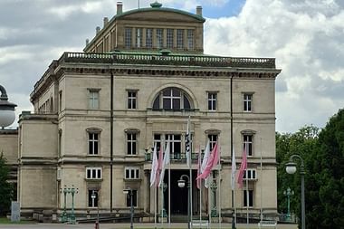 Villa Hügel, ein neoklassizistisches Herrenhaus in Essen mit cremefarbener Fassade, grünem Kupferdach und rosa Flaggen am Eingang unter bewölktem Himmel.