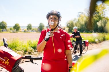 Radfahrer in rotem Shirt und Helm genießt Eis auf ländlichem Weg bei Höxter, mit anderem Radfahrer im Hintergrund und grünen Feldern.