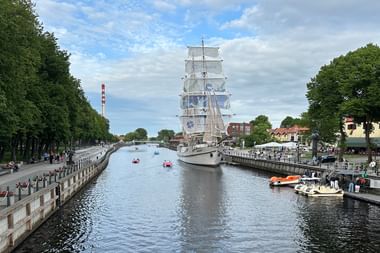 Weißer Großsegler mit vollen Segeln auf der Danė in Klaipėda, umgeben von baumgesäumten Promenaden, kleinen Booten und historischen Gebäuden.