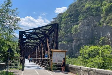 Schwarze Stahlfachwerkbrücke mit Radfahrer am Alpe Adria Radweg. Felsige Klippe mit grüner Vegetation erhebt sich hinter der Brücke unter blauem Himmel.