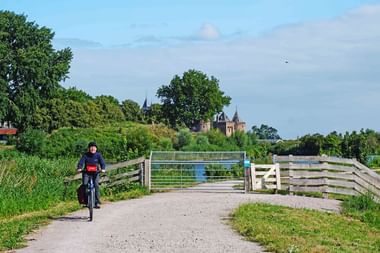 Radfahrer auf Schotterweg nähert sich Holztor mit Schloss Muiden im Hintergrund. Grüne Bäume und blauer Himmel rahmen die IJsselmeer-Route.