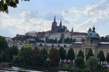 Prager Burg mit Veitsdom auf einem Hügel über historischen Gebäuden. Die Moldau fließt im Vordergrund unter blauem Himmel.