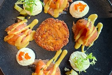 Kunstvoll angerichtete Vorspeise auf schwarzem Schiefer mit goldener Krokette in der Mitte, umgeben von Räucherlachsröllchen mit gelber Sauce.