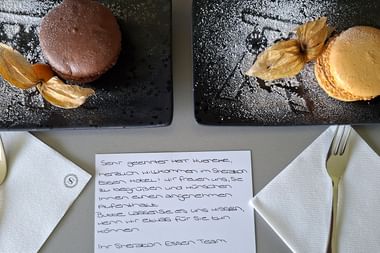 Zwei Dessertteller mit Macarons und Physalis auf schwarzem Schiefer, begleitet von einer handgeschriebenen Willkommensnachricht vom Sheraton Essen Team.