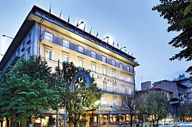 Mehrstöckiges Hotelgebäude mit gelber Fassade und beleuchteten Fenstern in der Dämmerung, umgeben von Bäumen und Außensitzbereich.