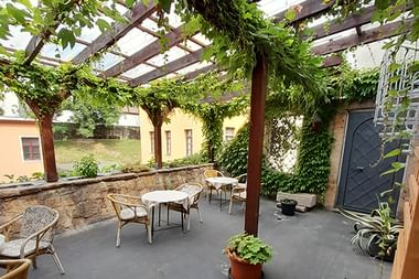 Überdachte Terrasse mit weinbewachsener Pergola in der Schlossschänke Bautzen. Steinmauern, Bistrotische und Stühle auf grauem Boden.