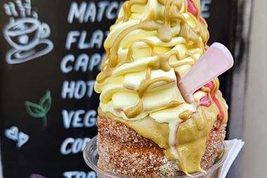 Traditionelles tschechisches Trdelník-Gebäck gefüllt mit Vanille-Softeis und Karamellsauce, vor einer Café-Menütafel gehalten.