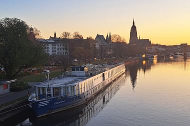 Flusskreuzfahrtschiff MS Casanova am Morgen mit historischer Stadtsilhouette und Kirchtürmen, die sich im ruhigen Wasser spiegeln.