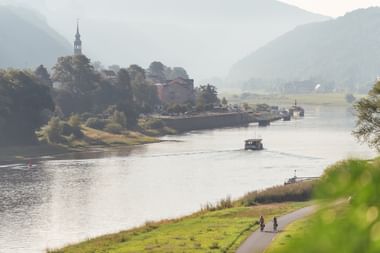 Scenic view of the Elbe River with cyclists on the riverside path. A historic town with church spire sits on the opposite bank, surrounded by hills.