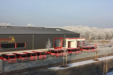 Luftaufnahme der Rückenwind-Anlage mit grauem Industriegebäude und Flotte roter Busse davor. Bereiftes Bäume umgeben das Gelände.