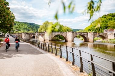 Zwei Radfahrer auf dem Weserradweg in Hann. Münden mit historischer Steinbrücke im Hintergrund und grünen Hügeln dahinter.