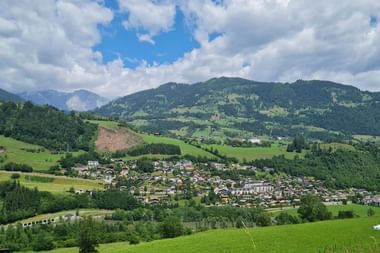 Panoramablick auf ein Dorf in einem grünen Alpental mit bewaldeten Hügeln und Bergen unter blauem Himmel mit weißen Wolken.
