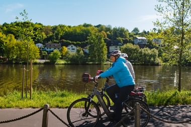 Zwei Radfahrer mit Helmen machen Pause an der Weser im Weserbergland. Häuser und grüne Hügel sind jenseits des Wassers unter blauem Himmel sichtbar.