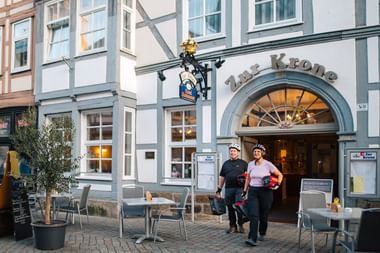 Zwei Radfahrer mit Helmen stehen vor dem Restaurant Zur Krone in Hamelns historischer Altstadt mit traditionellen Fachwerkhäusern.