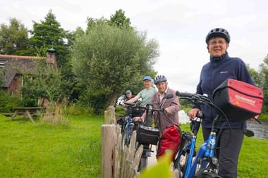 Gruppe von Radfahrern mit Helmen und Rädern auf grasigem Kanalweg. Rote Rückenwind-Tasche am Lenker sichtbar. Altes Backsteingebäude und Bäume im Hintergrund.