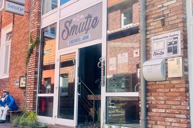 Backsteingebäude mit Eingang zum Hotel Smutje. Weißes Schild zeigt 'Stadthotel-GmbH'. Zwei Personen sitzen auf einer Bank vor dem Eingang.