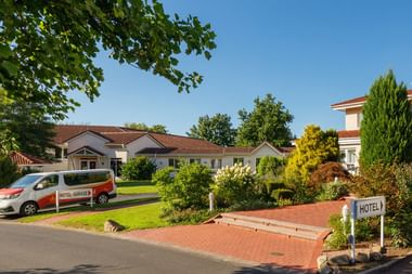 Weiße Hotelgebäude mit roten Ziegeldächern in Auefeld, umgeben von grünem Rasen und buntem Garten. Hotel-Shuttle vor dem Gebäude.