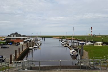 Kleiner Hafen mit Segelbooten an Holzstegen. Links ein Backsteingebäude und Parkplatz, rechts grünes Marschland.