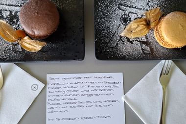 Zwei Schieferplatten mit Macarons und Physalis auf Puderzucker, daneben Servietten, Gabeln und eine handgeschriebene Willkommensnachricht vom Sheraton Essen.