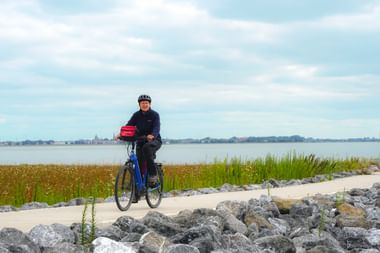 Radfahrer auf Weg am IJsselmeer mit felsigem Ufer, Schilf und entfernter Stadtsilhouette unter bewölktem Himmel.