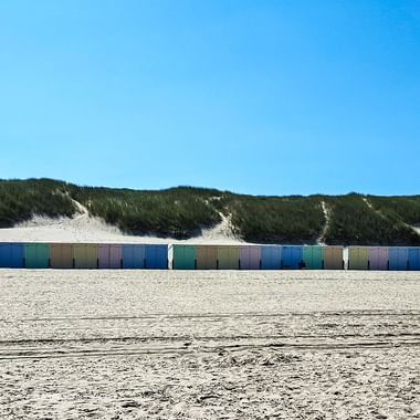 Reihe bunter Strandhäuser entlang des Sandstrandes in Oostkapelle, Zeeland. Grüne Dünen und klarer blauer Himmel im Hintergrund.