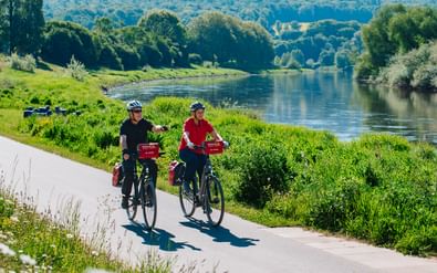 Zwei Radfahrer mit Helmen und Packtaschen fahren auf befestigtem Weg entlang der Weser. Grüne Wiesen und Bäume säumen das Flussufer bei Sonnenschein.