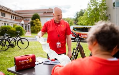 Kahlköpfiger Mann im roten Rückenwind-Shirt mit Ausweis erklärt Routenkarte einer sitzenden Person. Fahrräder und rote Ausrüstungstasche im Hintergrund.