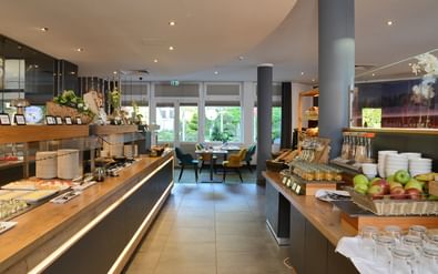 Moderner Hotel-Frühstücksraum mit Holz-Buffettheken, frischem Obst, Gebäck und Getränken. Graue Säulen und Sitzbereich im Hintergrund.