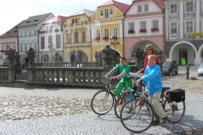 Drei Radfahrer mit Rädern posieren an einem verzierten Brunnen auf dem historischen Marktplatz von Kadan, umgeben von bunten Barockhäusern.