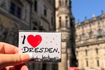 Hand hält einen 'I Love Dresden' Souvenirmagneten mit rotem Herz und Stadtsilhouette vor historischen Dresdner Gebäuden.