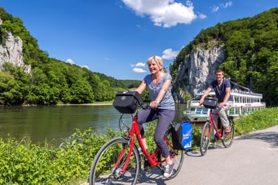 Zwei Radfahrer mit roten Rädern pausieren am Flussufer beim Weltenburg Donaudurchbruch. Weiße Kalksteinfelsen und Passagierschiff auf Donau.