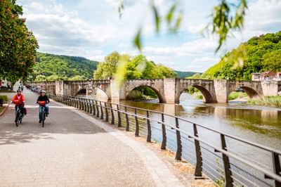 Zwei Radfahrer auf dem Weserweg in Hann. Münden mit historischer Steinbogenbrücke und grünen Hügeln im Hintergrund.