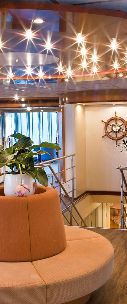 Modern reception area on SE Manon ship with curved wooden counter, circular seating, decorative ceiling lights, and nautical wheel decoration.