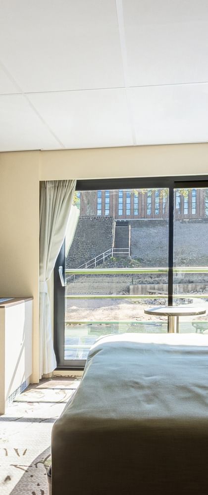 Modern ship cabin with double bed, wall-mounted TV, desk area, and large window with river view. White and brown interior design.