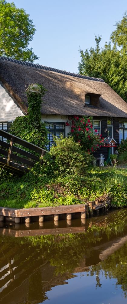 Traditionelles Reetdachhaus neben einem Kanal in Giethoorn mit Holzbrücke und Booten am Ufer, umgeben von üppigem Grün.