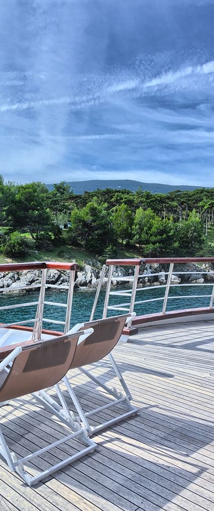 Bug-Sonnendeck der MY Andela Lora mit Holzdeck, Liegestühlen, Metallgeländer und Blick auf türkisfarbenes Wasser und grüne Küste.