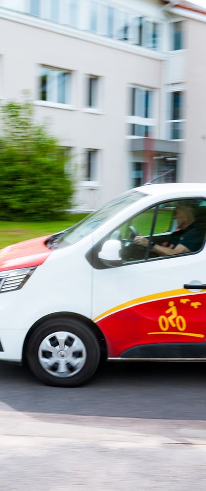 Weißer und roter Rückenwind-Van mit Fahrrad-Logo fährt auf einer Straße an weißen Gebäuden und grünen Bäumen vorbei in Bewegungsunschärfe.