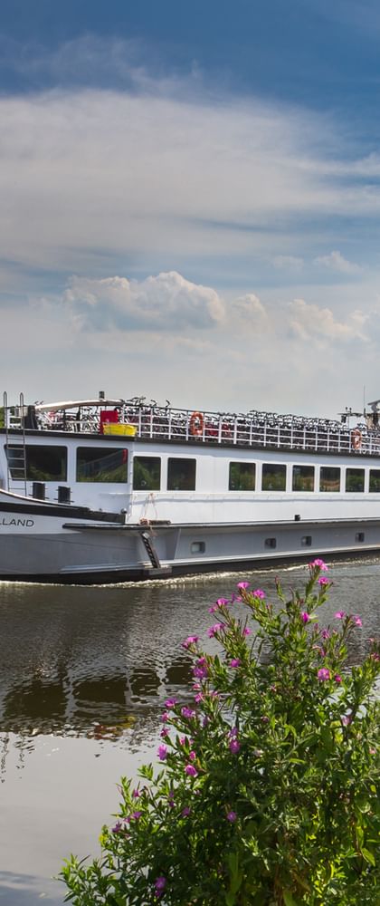 White river cruise ship De Holland sailing on calm water with wooden dock posts in foreground and flowering plants along the riverbank.