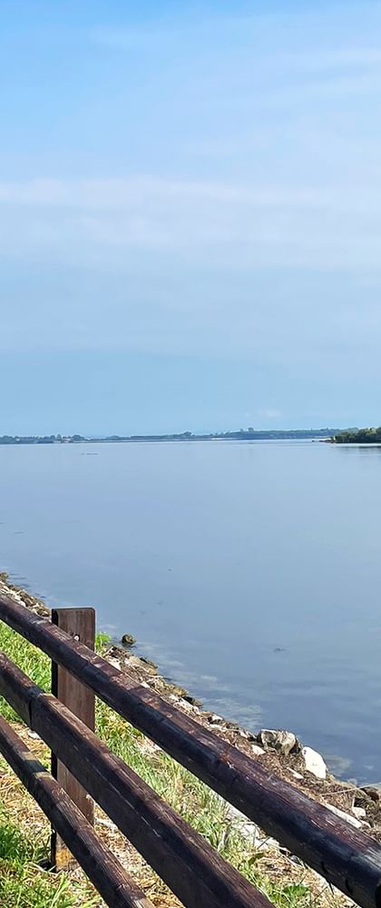 Zwei Radfahrer auf einer Brücke mit Holzgeländer am Alpe Adria Radweg bei Grado, mit ruhiger Lagune und ferner Küstenlinie unter blauem Himmel.