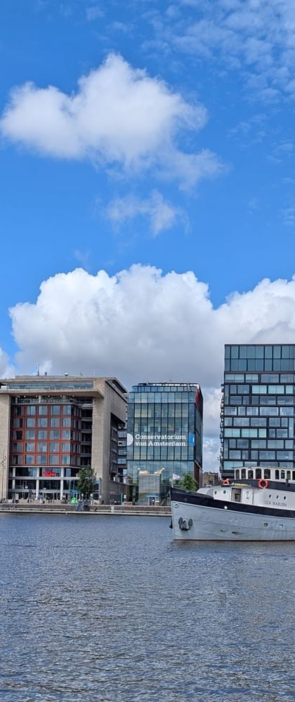 Weißes Passagierschiff Floor van Aemstel am modernen Ufer mit zeitgenössischen Glas- und Betongebäuden unter blauem bewölktem Himmel.