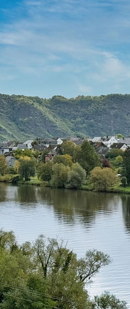 MS Olympia Flusskreuzfahrtschiff auf ruhigem Fluss mit Dorf und weinbergbedeckten Hügeln im Hintergrund unter blauem Himmel.