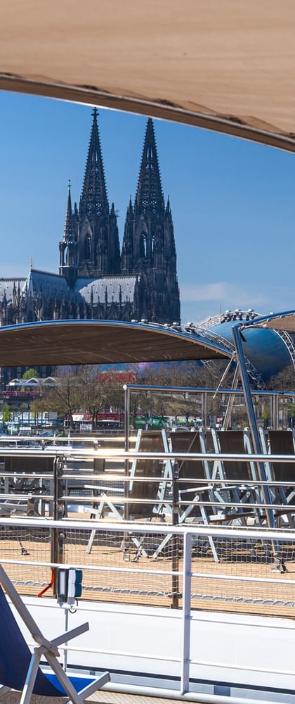 Sun deck of MS Lisabelle with blue deck chairs and red life ring, overlooking Cologne Cathedral's twin spires across the Rhine River.